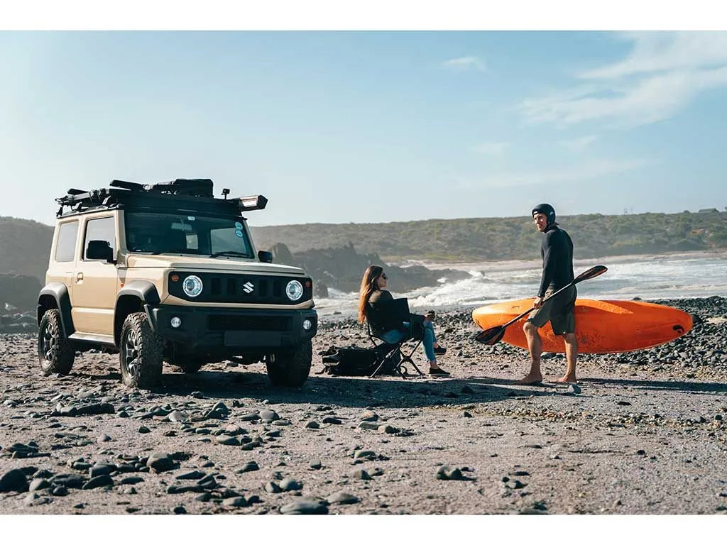 Beige SUV on a rocky beach with a person holding a kayak and another sitting by the vehicle.