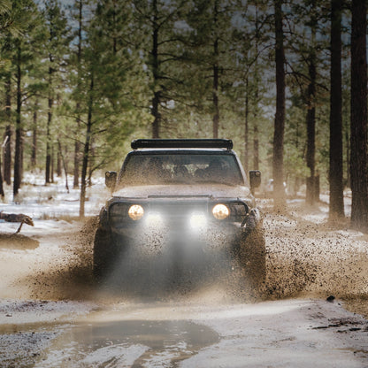 Jeep driving through a muddy forest road with trees in the background and using Noxsolis 9" LED Driving Lights - Spot Beam - 21,000 Lumens (Pair)